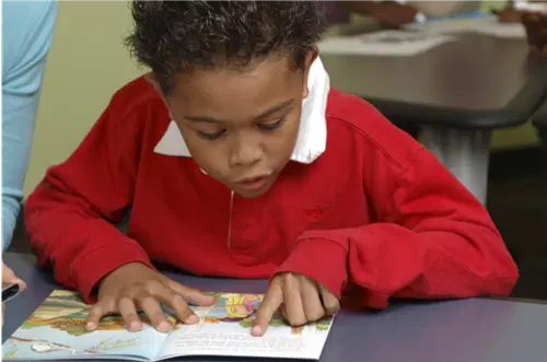 A child in a red shirt sits at a table and reads a picture book, using a finger to follow the text.