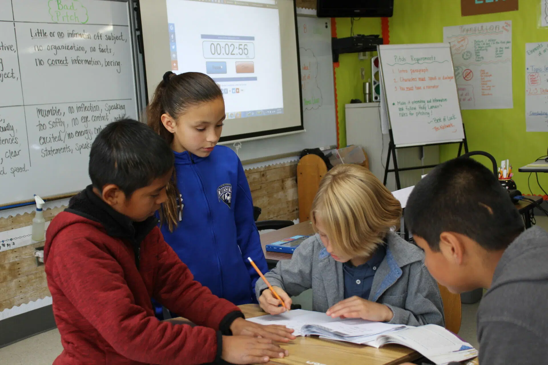 Four students work together at a desk in a classroom, with one writing in a notebook. A projector screen and whiteboards with notes are visible in the background.