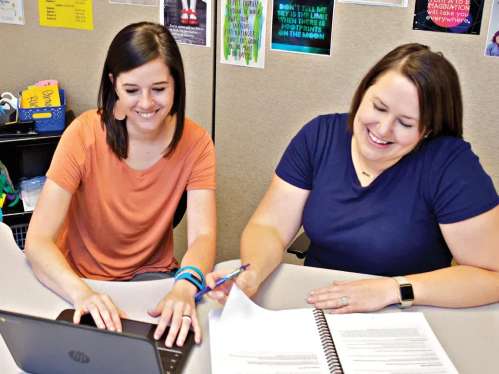 Two women are seated at a desk, smiling and working together on a project with a laptop and notebook in an office setting.