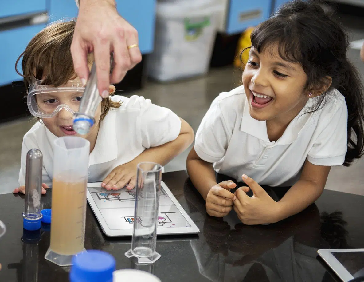 Two children in a lab setting engage in a science experiment with test tubes and a tablet. One child wears safety goggles, while an adult’s hand assists.