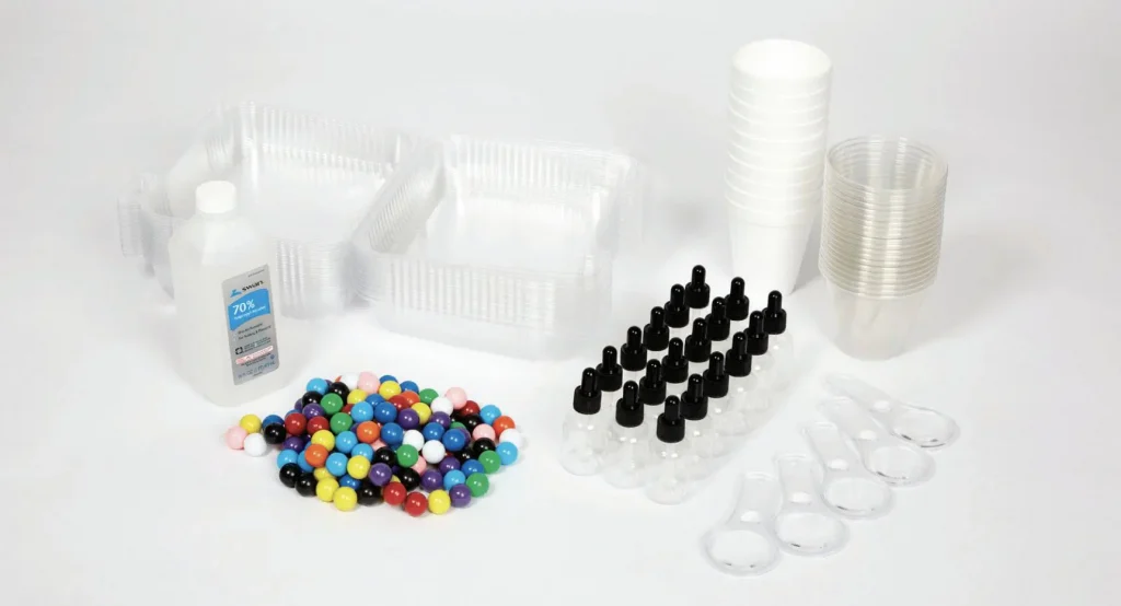A collection of clear plastic containers, empty dropper bottles, plastic cups, measuring spoons, a bottle of isopropyl alcohol, and colorful gumballs arranged on a white background.