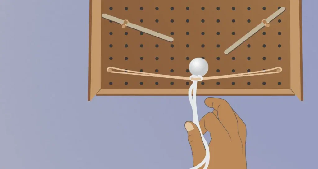 A hand pulls a white rope looped around a pegboard with angled dowels and a central white ball.
