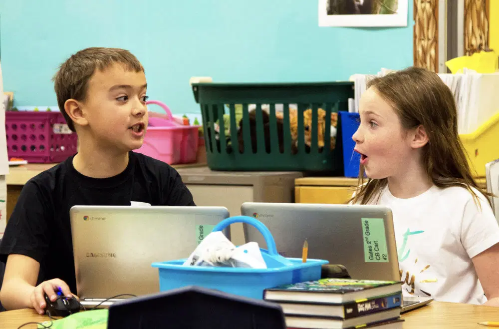 Two children sit at a table with Chromebooks, facing each other and talking, in a classroom with books and storage bins in the background.