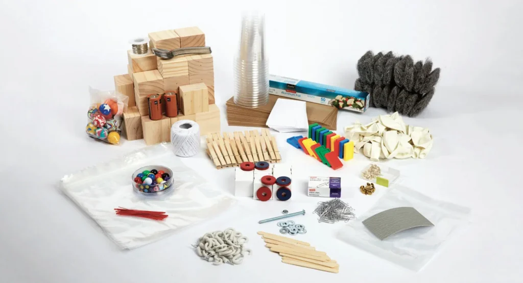 Assorted crafting supplies including wooden blocks, balloons, cups, string, clothespins, rubber bands, dominos, marbles, pipe cleaners, and various hardware items on a white background.