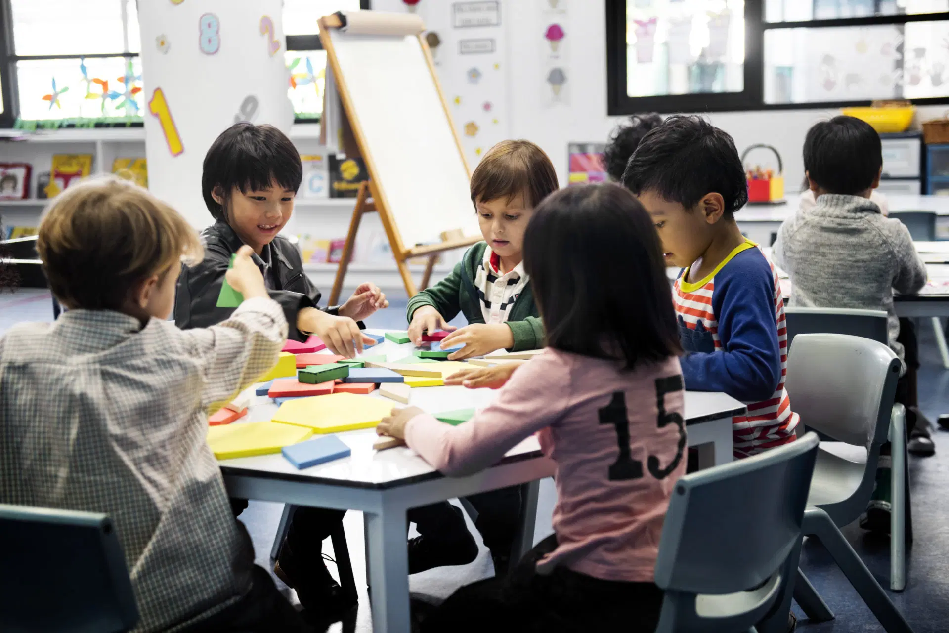 A group of young children sit around a table in a classroom, engaging in an activity with colorful geometric blocks and paper.