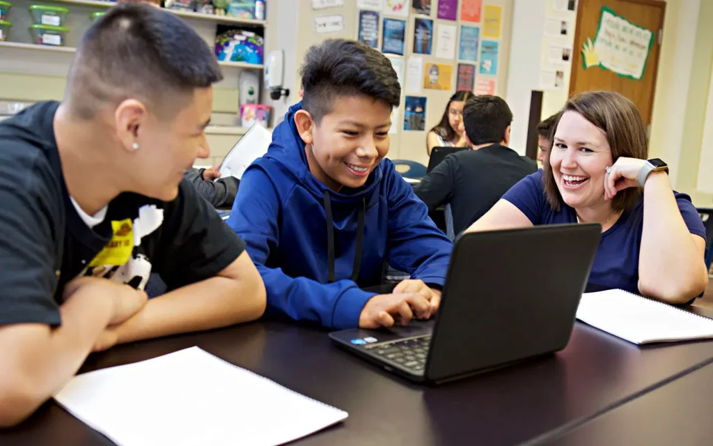 Three students sit at a table in a classroom, smiling and looking at a laptop screen with notebooks open in front of them.