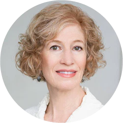 A woman with short, curly blonde hair and light skin wears a white top and earrings, smiling softly at the camera against a neutral background.