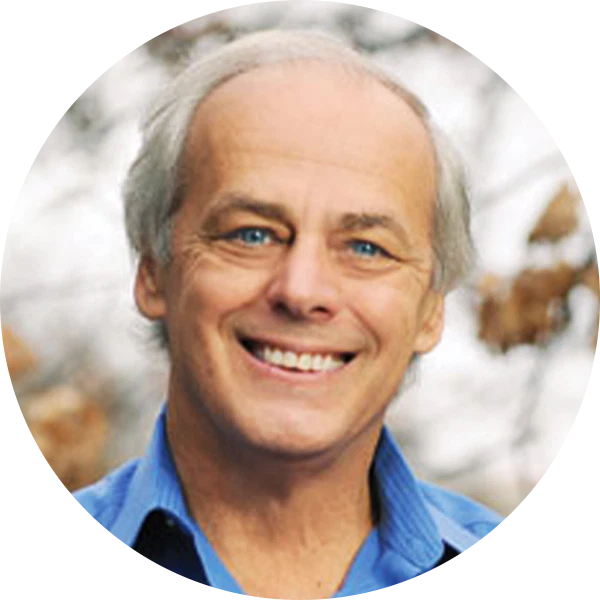 A smiling older man with light skin, gray hair, and blue eyes is wearing a blue collared shirt. The background is blurred with autumn leaves visible.