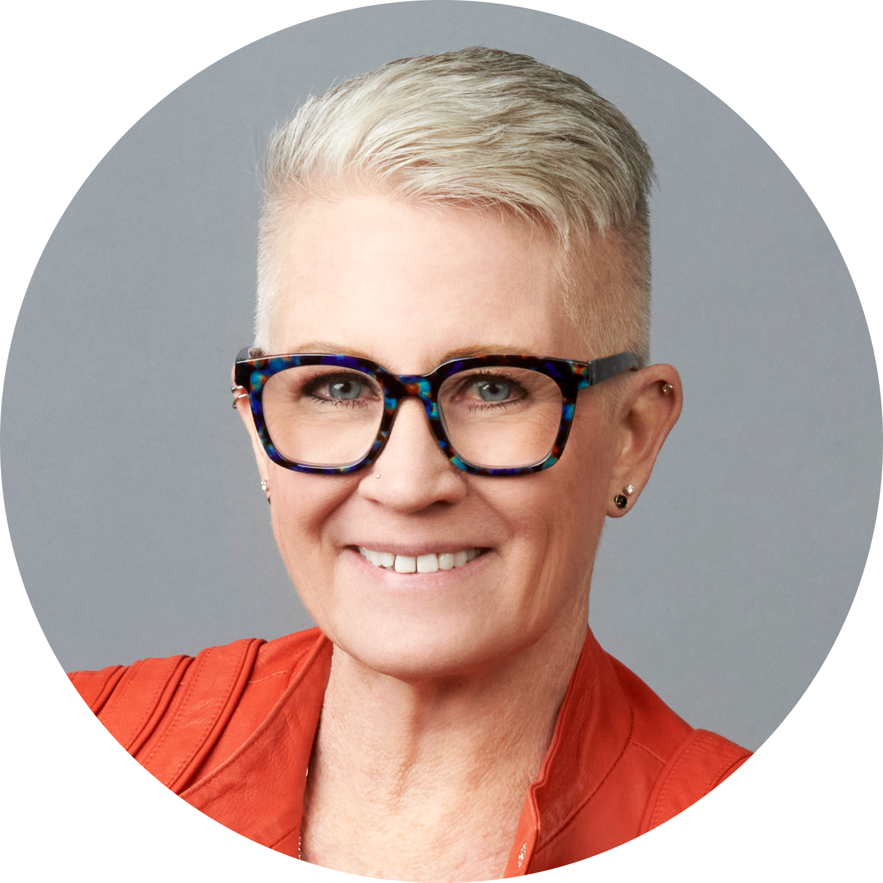 A person with short, light-colored hair, wearing dark-rimmed glasses, a red jacket, and small earrings, smiling at the camera against a neutral background.