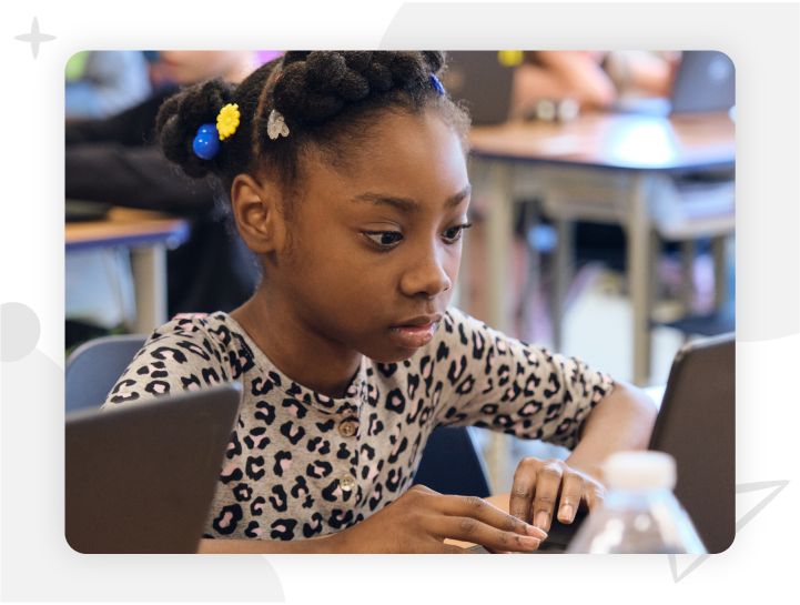 A young girl in a leopard print shirt works on a laptop at a classroom desk, with other students and desks visible in the background.