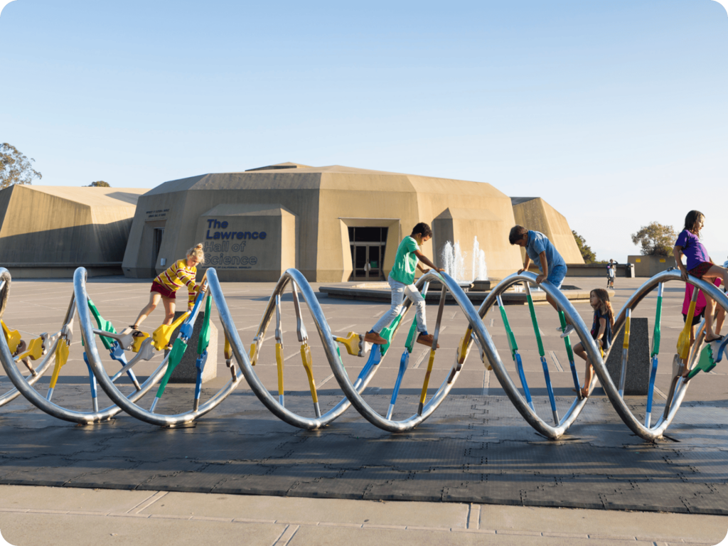 Children climb on a large DNA double helix sculpture outside The Lawrence Hall of Science, with the building and fountains visible in the background.