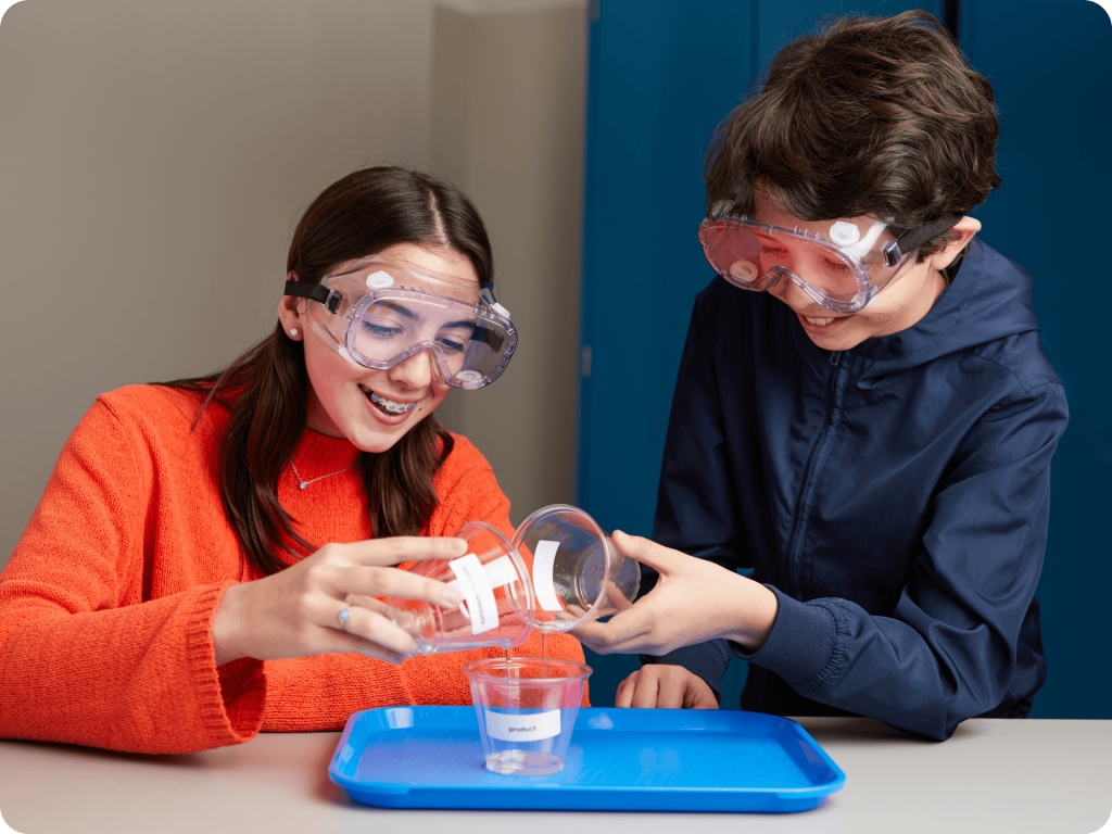 Two children wearing safety goggles conduct a science experiment, pouring liquid from one beaker into another over a blue tray on a table.
