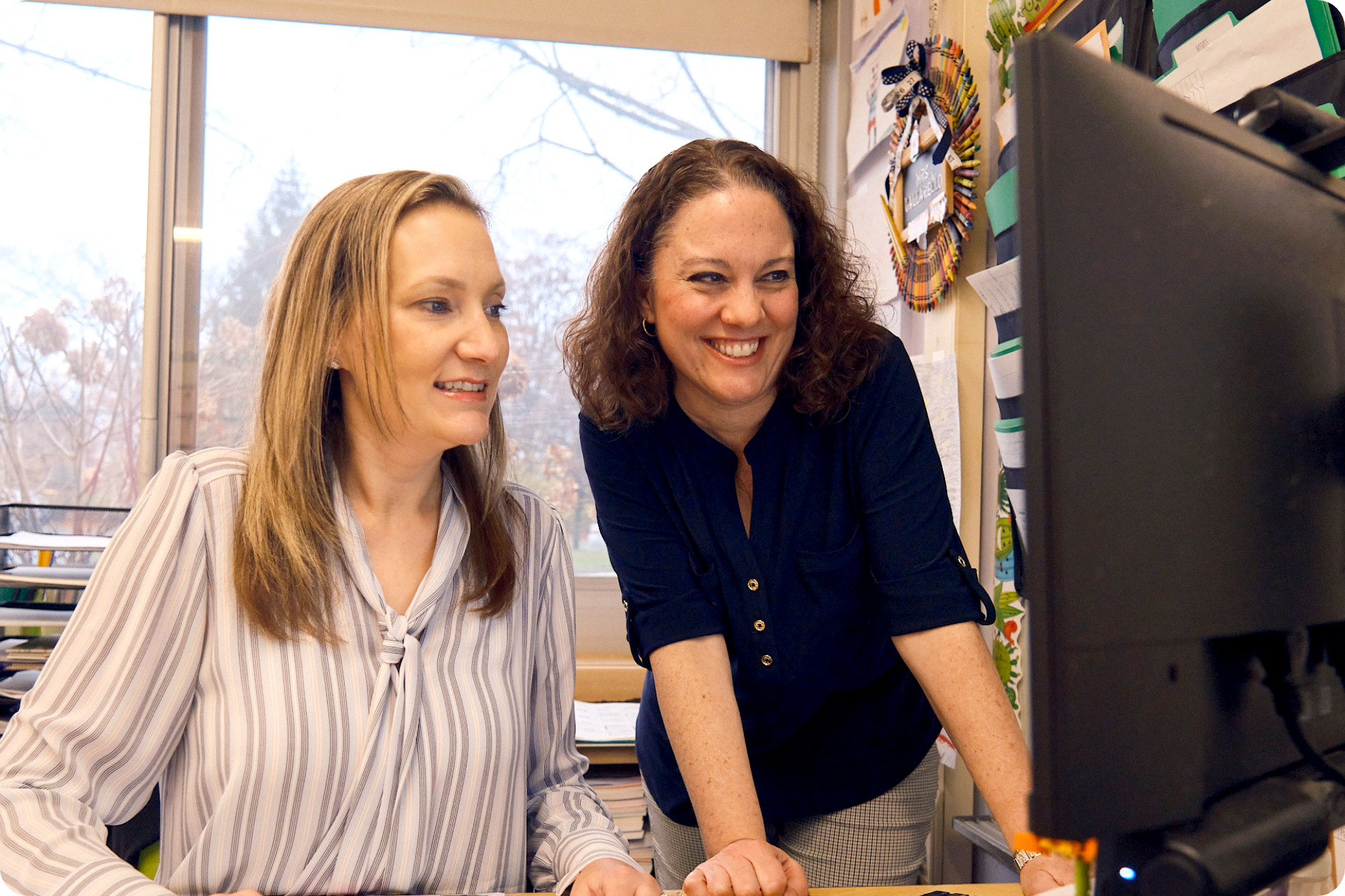 Two women in an office smile while looking at a computer monitor, discussing early literacy strategies. Stacks of papers and decorations are visible in the background.