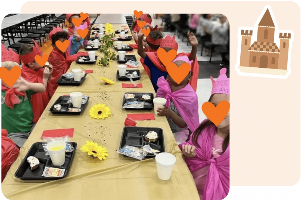 Children wearing red capes and paper crowns sit at a long, decorated table in a cafeteria, eating food from black trays; a castle graphic appears in the corner.
