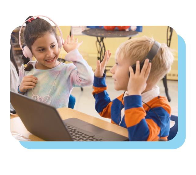 Two young children wearing headphones sit at a table with a laptop, smiling and giving each other high fives.