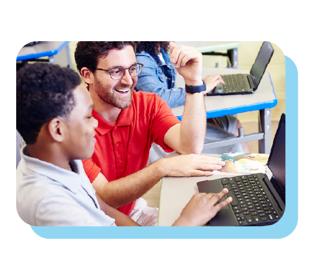 A teacher in a red shirt assists a student at a desk with a laptop in a classroom setting.