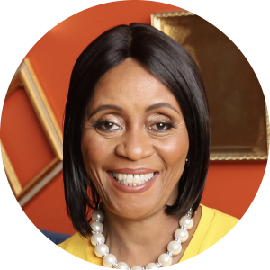 A woman with straight black hair and a pearl necklace is smiling in front of an orange wall with gold picture frames, ready to share educational insights on her teaching podcast.