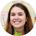 A young woman with long brown hair smiles at the camera, wearing hoop earrings and a lanyard, with colorful posters on the wall behind her.