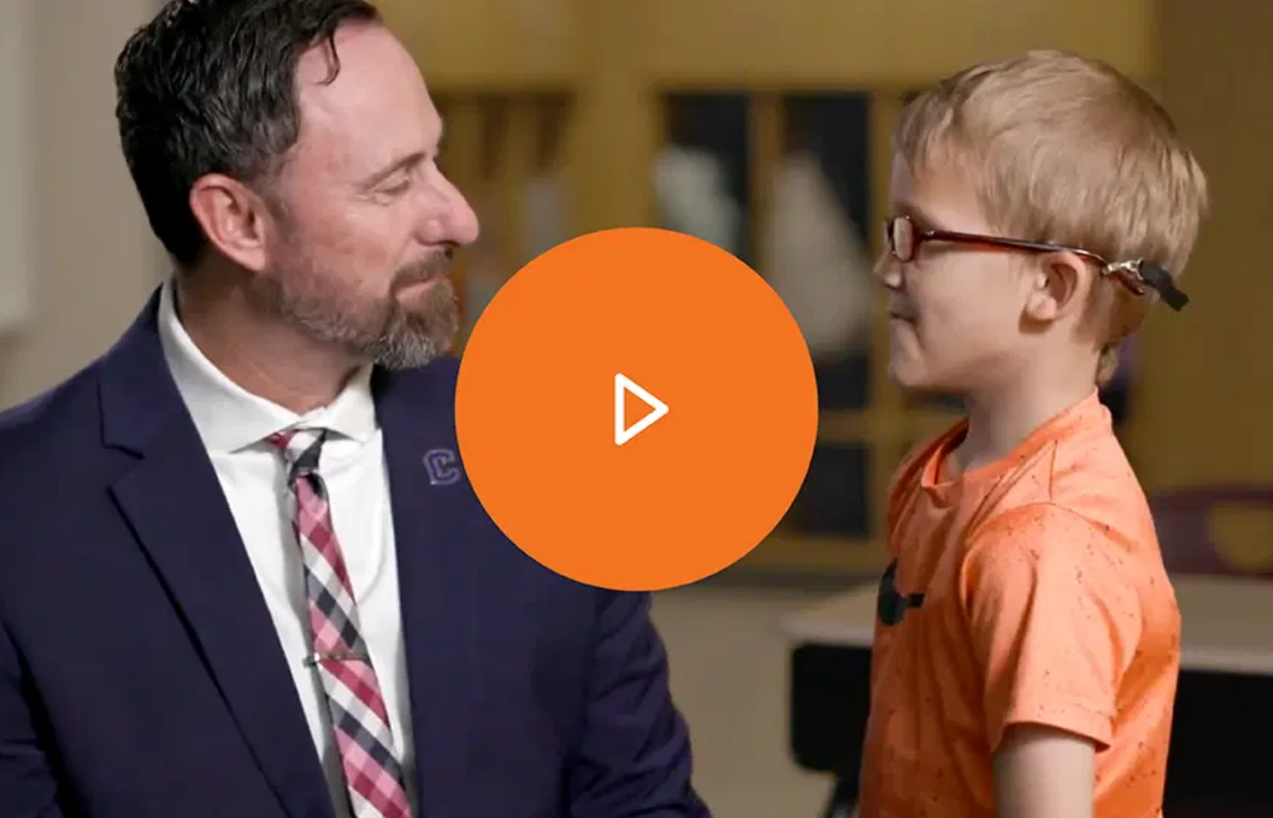 An adult man in a suit and a child with glasses and a hearing device smile at each other. An orange play button is centered over the image, highlighting k-5 teaching resources inspired by the science of reading.