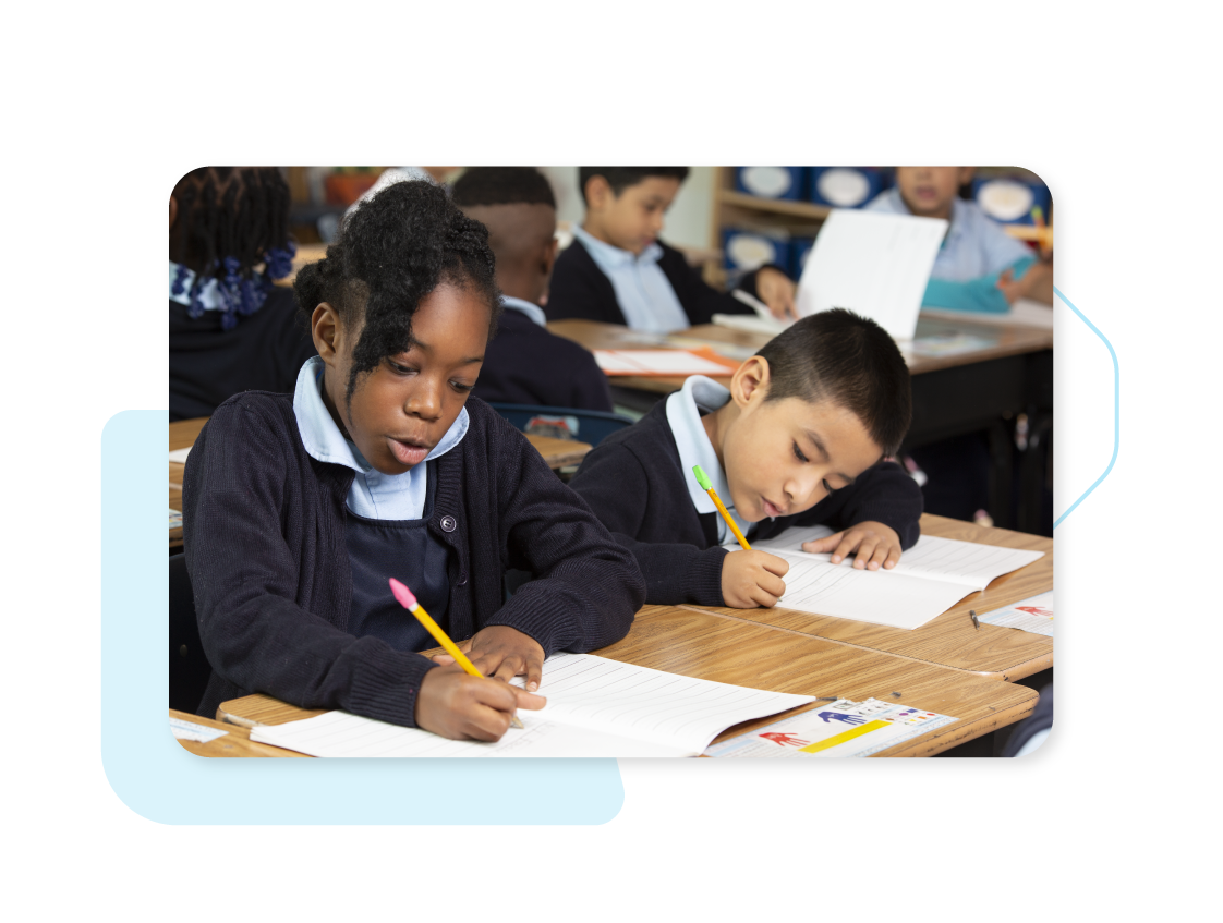 Two young students sitting at desks in a classroom, focused on writing in notebooks with pencils. Other students are visible in the background.
