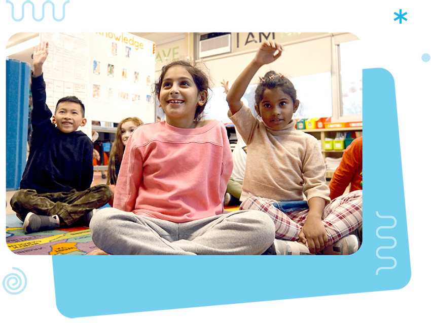Four young children sit on a classroom floor, two with their hands raised, participating in an activity. Shelves of books and educational posters highlight the classroom’s focus on bilingual programs in the background.