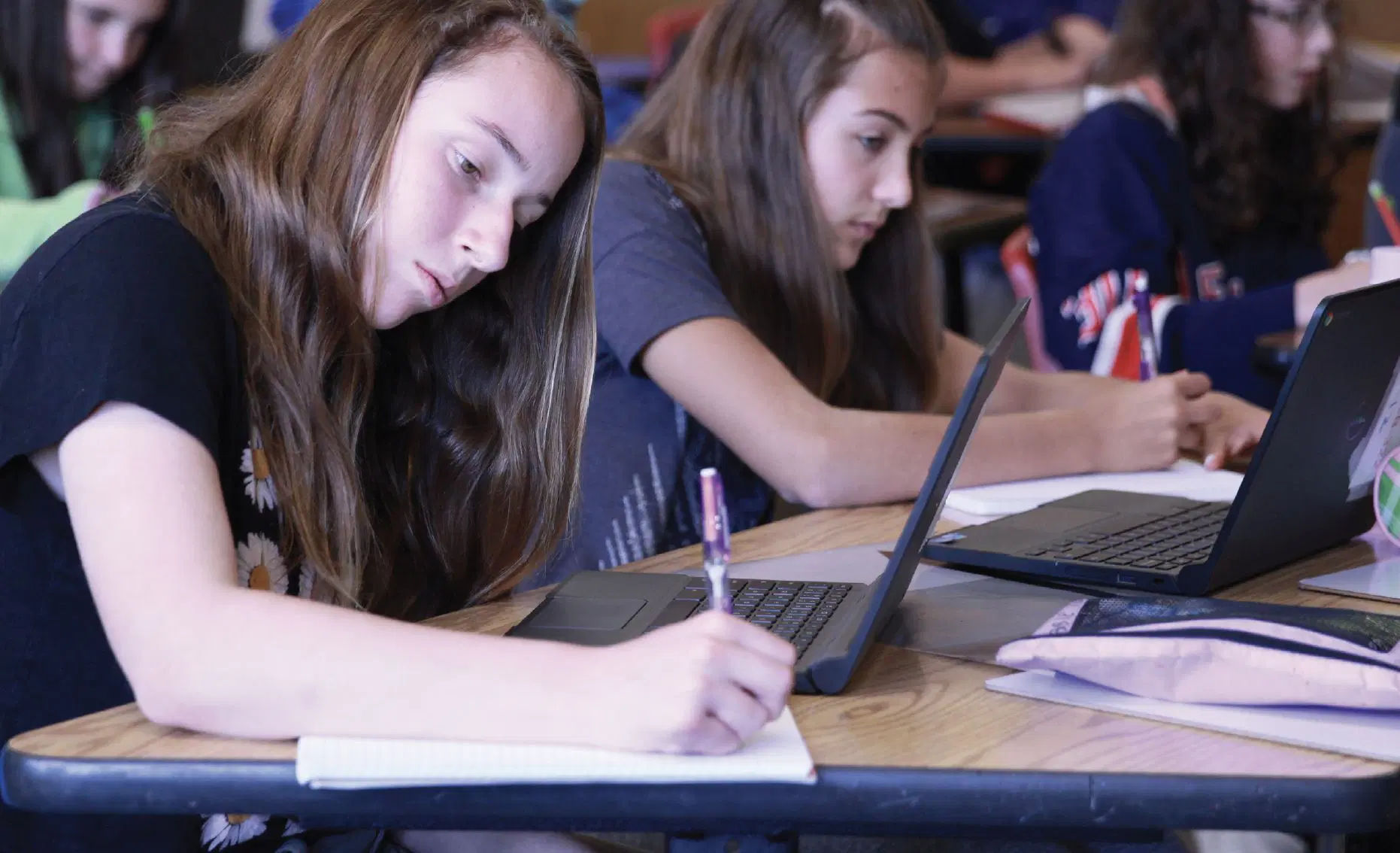 Two students sit at desks writing in notebooks and using laptops in a classroom setting.