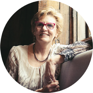 A woman with short blonde hair and red-framed glasses wearing a patterned blouse sits and smiles near a wooden wall.