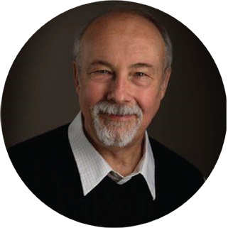 A man with a gray beard and mustache, wearing a white collared shirt and dark sweater, smiles while looking at the camera.