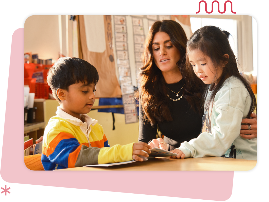 A teacher sits at a table with two young students, guiding them as they look at a book together in a classroom setting.