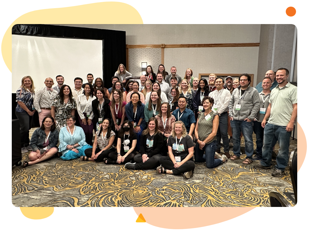 A large group of people, including math teachers, pose together indoors, some standing and others seated on the floor, with a projection screen in the background showcasing Desmos Classroom tools.