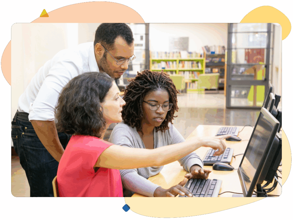 Three people working together at a row of computers in a library setting. One person is pointing at the screen.