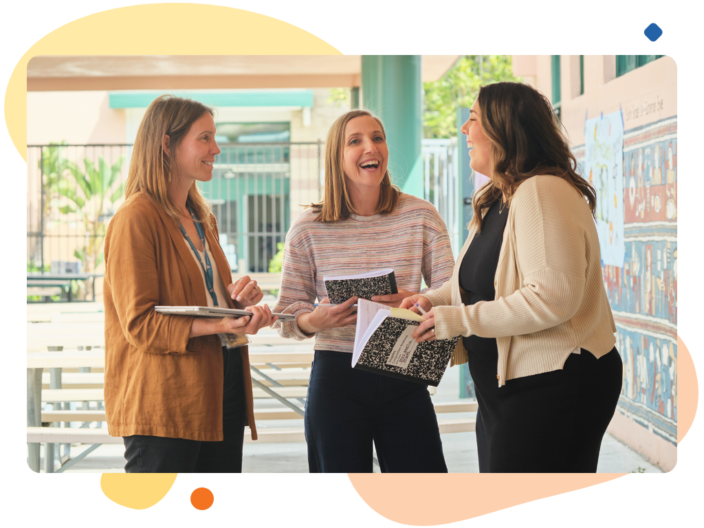 Three people stand outside a building, holding notebooks and papers. They are engaged in conversation about Desmos Classroom tools and appear to be smiling, likely sharing their passion for teaching math.