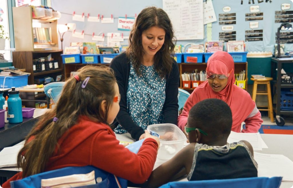 A teacher sits at a table with three young students in a classroom, engaging in an Amplify Science group activity with papers and a clear plastic container.