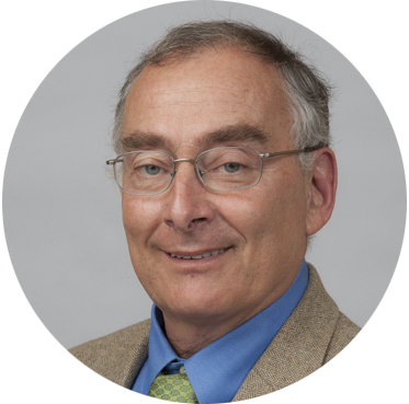 A man with glasses, short gray hair, and a receding hairline is wearing a blue shirt, green tie, and beige suit jacket, looking at the camera with a slight smile.