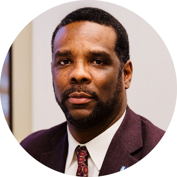 A man with short hair and a beard wearing a suit and tie is looking directly at the camera.