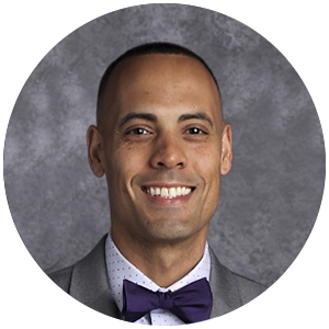 A man wearing a grey suit, white shirt with blue dots, and a purple bow tie smiling at the camera against a grey background.