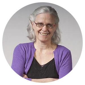 Individual with glasses and gray hair, wearing a purple cardigan and black top, smiling with arms crossed.