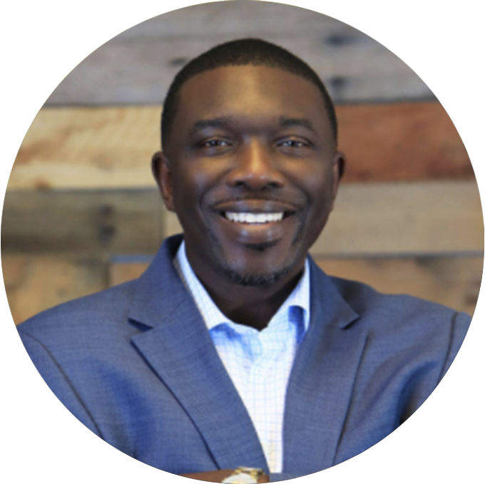 A man in a blue suit jacket and white shirt smiles at the camera, standing against a wooden background.