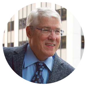 An older man with gray hair wearing glasses, a blue shirt, and a patterned tie is smiling while standing in front of a building with large windows.