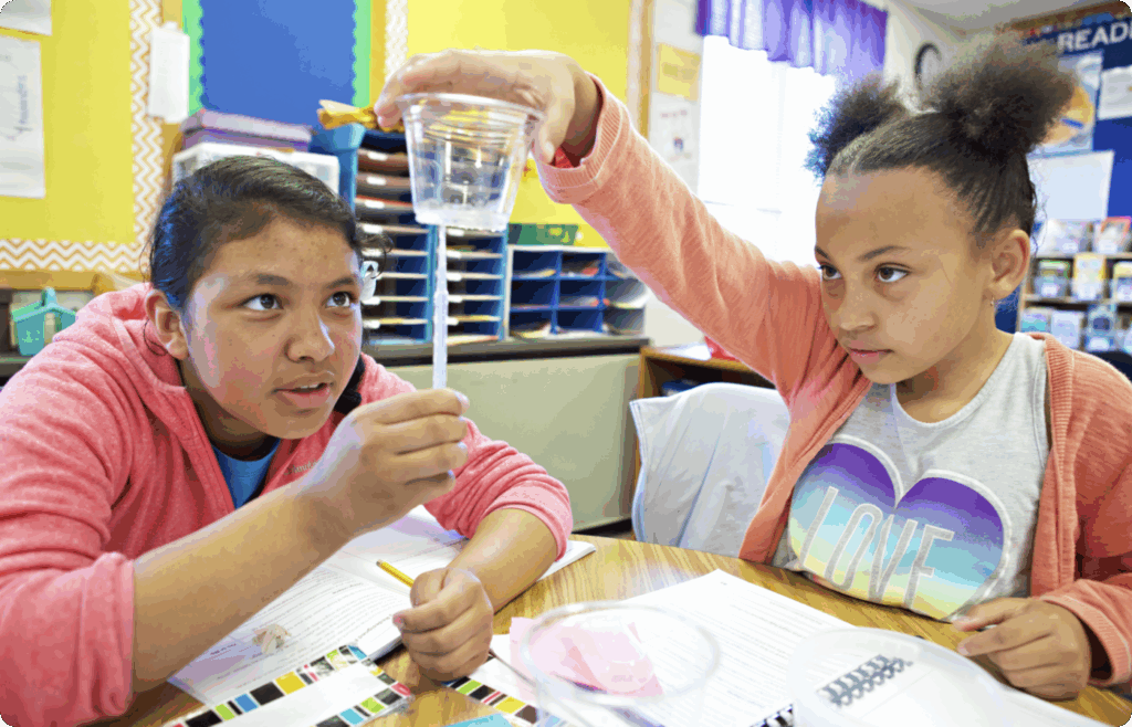 Two girls at a classroom desk conduct an NGSS-aligned science experiment, holding up a clear plastic cup with water and a straw, with papers and materials spread out on the table.