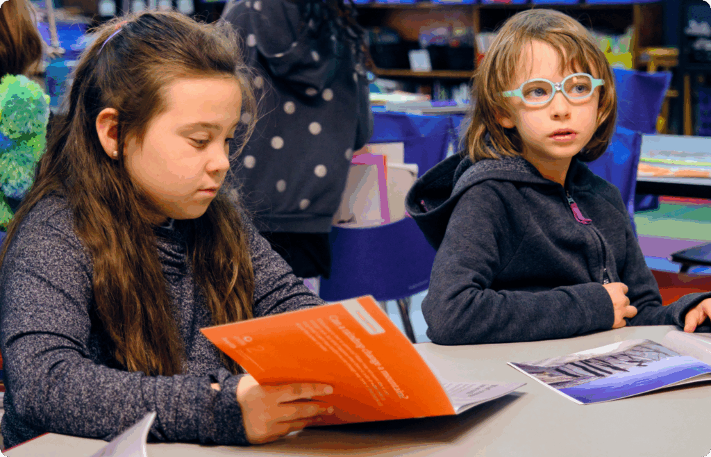 Two children sit at a classroom table; one reads an orange booklet, while the other looks ahead with an open magazine, both engaged in an NGSS-inspired activity.