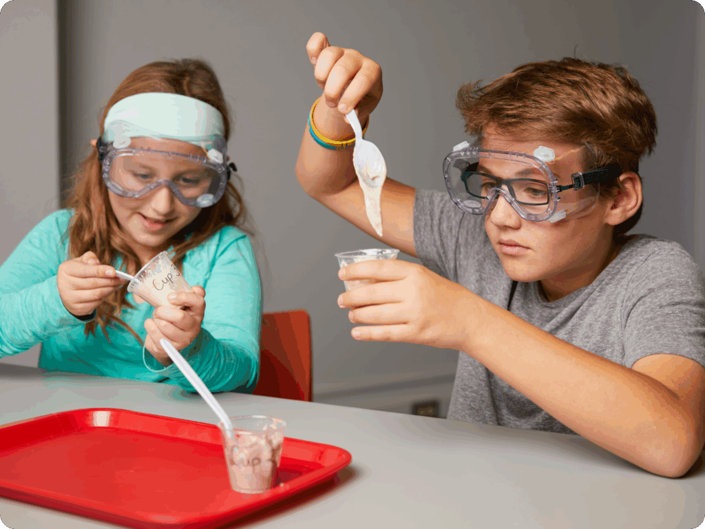Two children wearing safety goggles, following NGSS guidelines, mix substances in clear plastic cups using plastic spoons, seated at a table with a red tray.